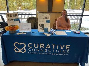 Curative Connections table at a event - featuring a blue tablecloth and a volunteer in a pink sweater.