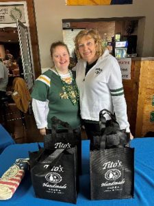 Two women wearing Green Bay Packer sweaters pose behind a trade table filled with tote bags.