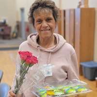 An older woman in a pink sweatshirt smiles for the camera as she holds a small bouquet of roses and a tray of cupcakes. 