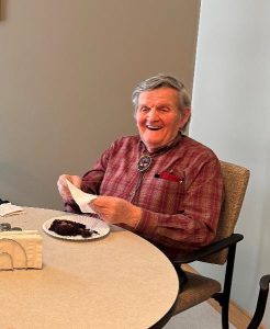 A gentleman in a red plaid shirt smiles as he eats a piece of chocolate cake.